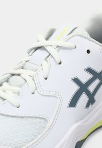 Hvit atletisk sko med en teksturert overdel, grå aksenter og gule detaljer. Har flate lisser og en polstret såledesign.