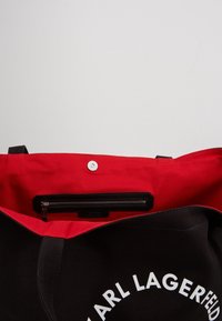 Černá taška přes rameno s červeným interiérem. Obsahuje kapsu na zip, magnetické zavírání a bílé logo textu vpředu. Vyrobena z odolné tkaniny.