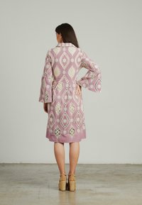 Abito lungo rosa a maniche lunghe con intricate fantasie bianche, dettagli in pizzo e vita cintrata. Presenta maniche svasate e un taglio fino al ginocchio.