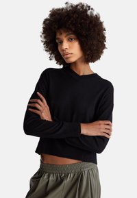 Kobieta o kręconych włosach, ubrana w czarny sweter z długim rękawem i dekoltem w serek oraz oliwkowozielone spodnie z elastycznym pasem, z założonymi skrzyżowanymi ramionami na białym tle.