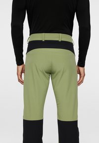 Miestne zelené výkonnostné nohavice s čiernym pásom a spodným panelom. Majú hladkú textúru a úzky strih pre lepšiu pohyblivosť.