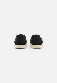 Schwarze Canvas-Slipper mit beigen Sohlen, versehen mit einem strukturierten Stoff und geprägtem Logo-Detail an der Ferse.