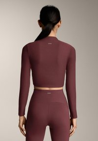 Bordeauxrode, geribbelde crop top met lange mouwen en bijpassende hoge taille legging. Subtiele logo-details op de bovenrug en tailleband.