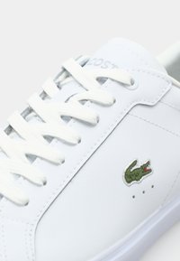 Biały skórzany sneaker z gumową podeszwą, z białymi sznurowadłami, delikatnym przeszyciem oraz zielonym wyszywanym logo krokodyla po boku.