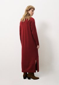 Robe tricotée longue et rouge avec des fentes sur les côtés, un col rond et des manches longues. Portée avec des bottes brunes à hauteur de cheville. Fond uni.