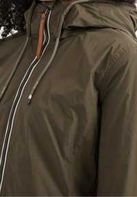Veste imperméable vert olive avec fermeture éclair, capuche ajustable et accents bruns contrastants. Présente une texture lisse et un tissu durable.