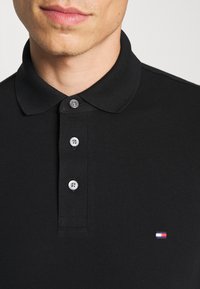 Crna polo majica s teksturiranim materijalom, tri siva gumba i mali izvezeni logo u crvenoj, bijeloj i plavoj boji na prsima.