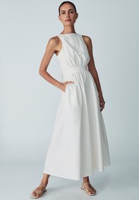 Weißes ärmelloses Kleid mit tailliertem Oberteil, elastischem Bund und Taschen. A-Linie Rock, der bis zum Knöchel fließt. Glattes Material, minimalistische Details.
