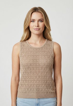 Mujer rubia con un top de punto sin mangas beige con un delicado patrón de encaje y jeans azul claro, de pie frente a un fondo liso.