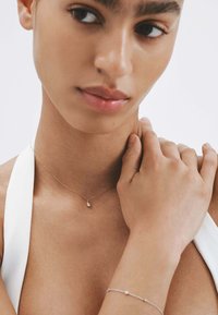 Collar de oro con un colgante en forma de lágrima, llevado por una modelo. Una pulsera de plata con una pequeña cuenta es visible en la muñeca. Textura de piel suave.