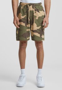 Camo shorts in green and tan, made of soft fabric, with an elastic waistband and drawstring. Paired with white sneakers and socks.