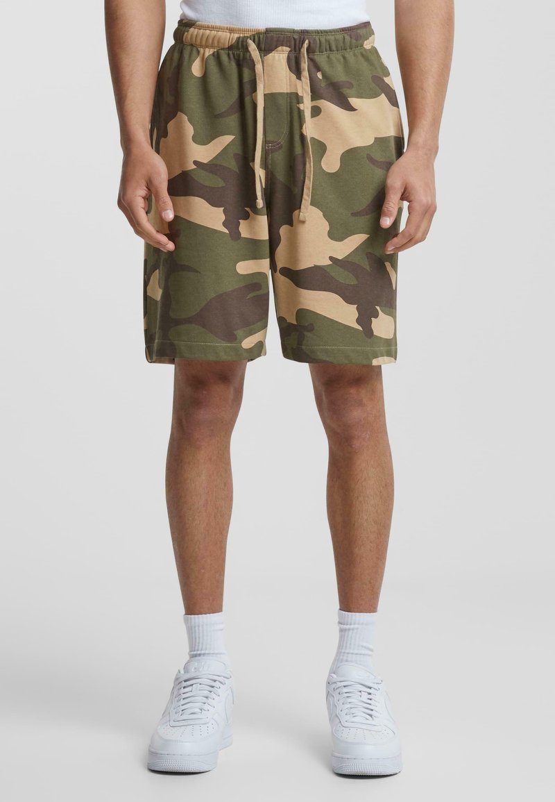 Camo shorts in green and tan, made of soft fabric, with an elastic waistband and drawstring. Paired with white sneakers and socks.