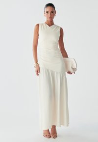 Vestido largo en color crema, con un corpiño ajustado de acabado texturizado y una parte inferior con vuelo. Combinado con un bolso de mano y sandalias con tacón.