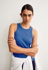 Blue ribbed tank top with a round neckline, featuring wide shoulder straps. A white shirt is tied casually around the waist.