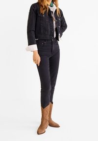 Mujer vistiendo una chaqueta de mezclilla negra corta, jeans negros de talle alto, puños de camisa blanca visibles y botas de vaquero de ante marrón, de pie sobre un fondo blanco.