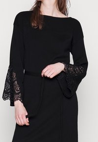 Černé šaty s dlouhými rozšířenými rukávy, které mají složité krajkové detaily. Vyrobené z měkkého materiálu, mají rovný siluetu a pásek v pase.