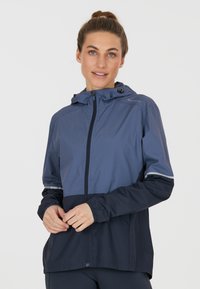 Giacca leggera e impermeabile in blu scuro e blu navy. Presenta una zip frontale, cappuccio, polsini elasticizzati e dettagli riflettenti sulle maniche.