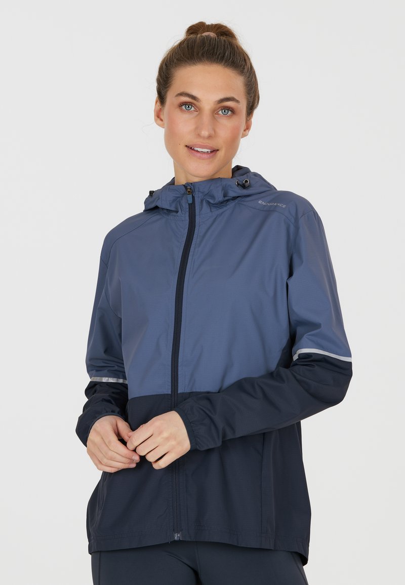 Giacca leggera e impermeabile in blu scuro e blu navy. Presenta una zip frontale, cappuccio, polsini elasticizzati e dettagli riflettenti sulle maniche.