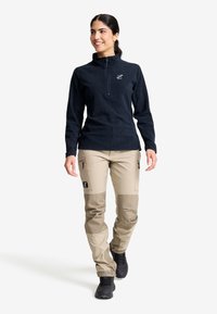 Frau geht, trägt einen schwarzen Fleece-Pullover mit Reißverschluss und beigefarbene Cargo-Hosen mit verstärkten Knie-Patches sowie schwarze Wanderschuhe.