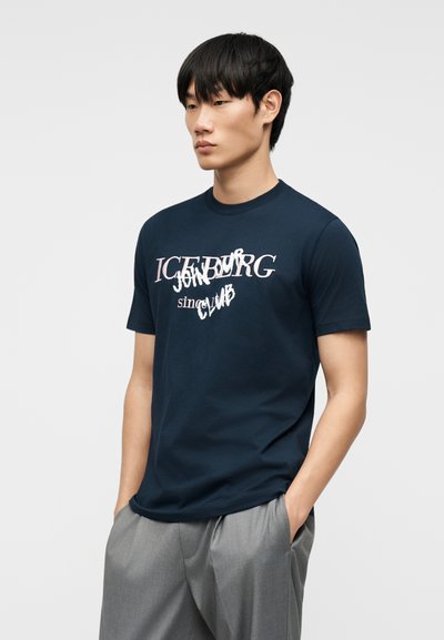 Νέος άνδρας με μαύρα μαλλιά που φοράει μπλε ναυτικό t-shirt με λευκό και ροζ κείμενο «ICENBURG Join Club», γκρι παντελόνι, χέρια στις τσέπες, ουδέτερο φόντο.
