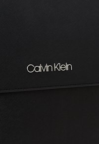 Surface en cuir noir texturé avec le logo de la marque "Calvin Klein" en métal argenté centré près du bord supérieur.