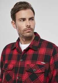Camisa de franela a cuadros rojos y negros con cierre de botones, dos bolsillos en el pecho y un pequeño acento de logo. Tejido suave con una apariencia texturizada.