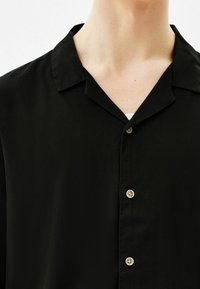 Camicetta nera a maniche corte con un tessuto morbido e texture, caratterizzata da un colletto alla coreana e due bottoni chiari lungo la patta.