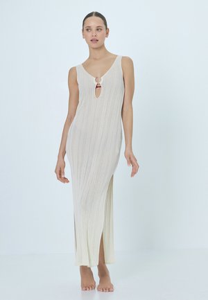 Frau in einem ärmellosen, cremefarbenen Strick-Maxikleid mit einem vorne angebrachten Ringdetail und seitlichen Schlitzen, barfuß vor einem schlichten Hintergrund stehend.