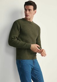 Sweter w kolorze oliwkowym, prążkowany, z długimi rękawami, okrągłym dekoltem i teksturowanym materiałem. Noszony z niebieskimi dżinsami. Prosty, dopasowany krój.