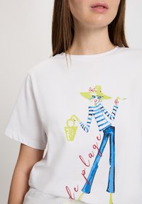 Biała bawełniana koszulka z kolorową grafiką przedstawiającą osobę w w paskowanej koszuli i kapeluszu przeciwsłonecznym, trzymającą torbę. Tekst brzmi "le plage."