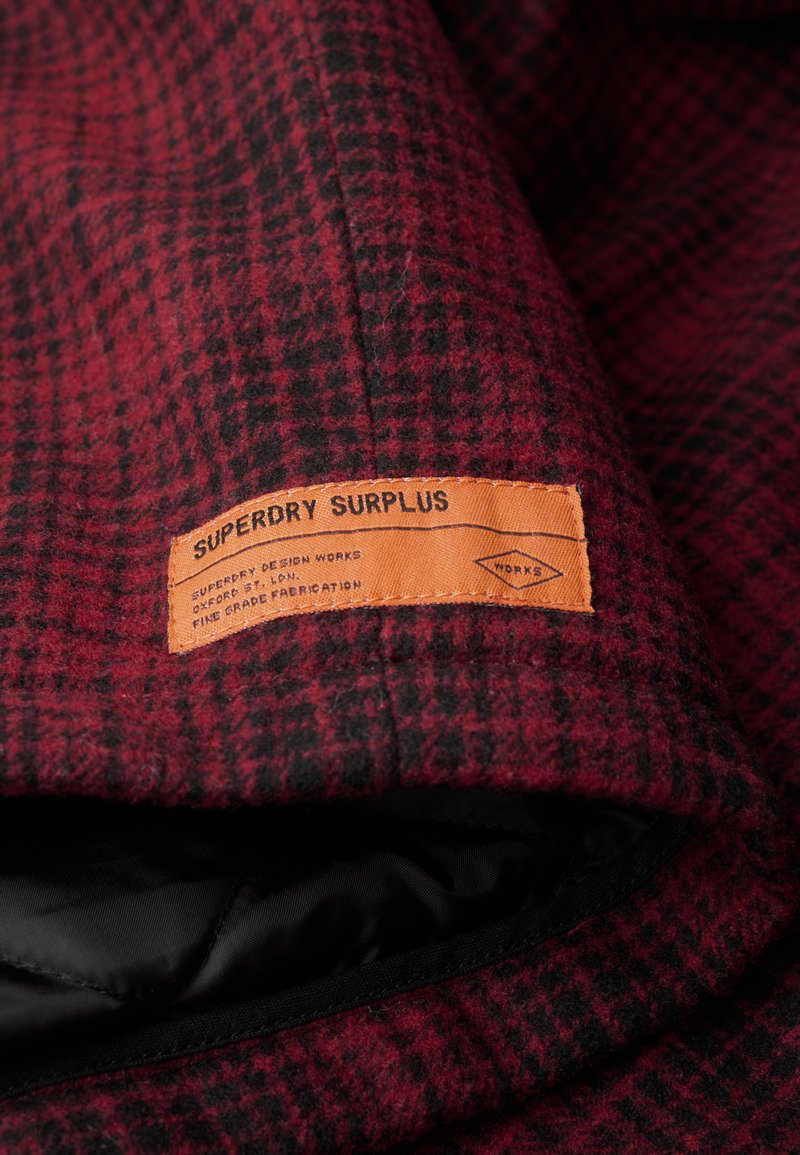 Rot-schwarz karierter Stoff mit grober Textur; ein orangefarbenes Etikett mit der Aufschrift "Superdry Surplus" ist sichtbar und gibt Marken- sowie Stoffdetails an.