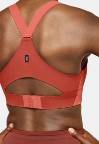 Sujetador deportivo de tejido rojo- anaranjado, con diseño de espalda nadadora, detalle recortado, cierre ajustable y textura suave.