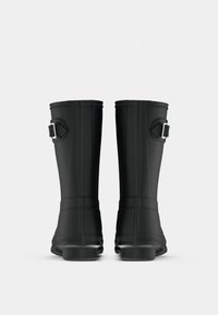 Μαύρες καουτσούκ γαλότσες διαθέτουν ψηλό σχέδιο, ομαλή υφή, μεταλλικό λουρί στον αστράγαλο και στιβαρή σόλα με λεπτομέρειες πρόσφυσης.