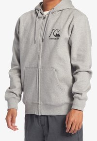 Sudadera con cierre en color gris, hecha de un suave forro polar. Presenta un logo en negro en la parte frontal, capucha con cordón y dos bolsillos laterales. Puños y bajo elásticos.