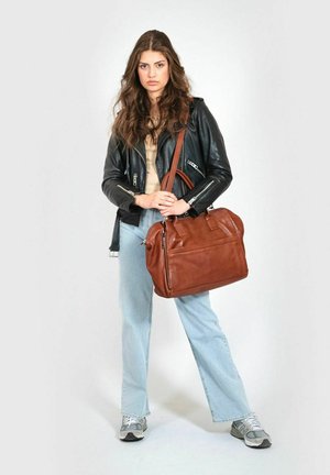 Brown leather handbag with dual handles and adjustable shoulder strap, featuring a front zip pocket, held by a person in a black leather jacket.