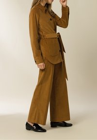 Ruskea corduroy-takki, jossa on vyötärönauha, nappikiinnitys ja läpätasku, yhdistettynä leveälahkeisiin samansävyisiin housuihin ja mustiin lakkaisiin kenkiin.