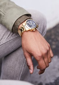Montre chronographe dorée avec un cadran bleu foncé, bracelet en métal et accents argentés. La montre est posée sur une surface texturée gris.