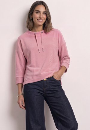 Femme aux cheveux bruns mi-longs souriant, portant un haut rose à manches longues avec des cordons et un jean bleu foncé, debout contre un mur clair.