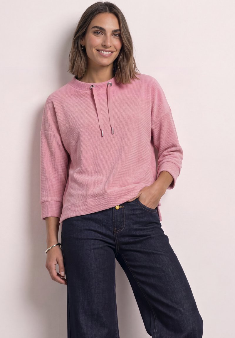 Femme aux cheveux bruns mi-longs souriant, portant un haut rose à manches longues avec des cordons et un jean bleu foncé, debout contre un mur clair.