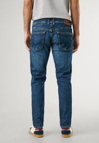 Donkerblauwe denim jeans met een slim fit, vijf zakken, zichtbare stiksels en een bruine leren patch op de tailleband. Casual schoeisel zichtbaar.