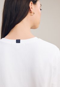 Femme aux longs cheveux foncés portant une chemise blanche avec une étiquette bleu marine au niveau de la nuque, et une boucle d’oreille créole dorée à l’oreille gauche.