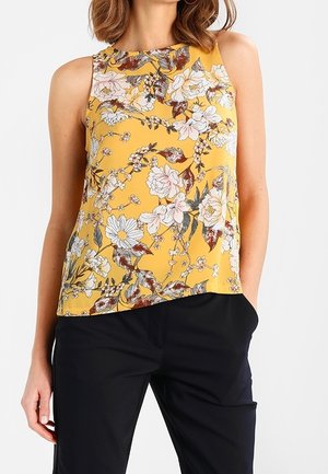 Vrouw draagt een mouwloos geel topje met wit en bruin bloemenpatroon en zwarte broek, staat met één hand in de zak.