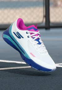 Weißer Sportschuh mit rosa und blauen Akzenten, strukturiertem Obermaterial und einer lebhaften blauen Sohle. Verfügt über ein kontrastierendes Logo und Schnürdesign.