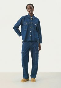 Conjunto de chaqueta de mezclilla y pantalones cargo en azul oscuro. La chaqueta cuenta con botones dorados y dos bolsillos frontales. Los pantalones tienen múltiples bolsillos y un corte holgado.