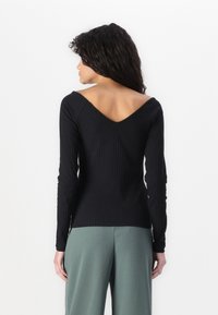 Black ribbed long-sleeve top with a deep V-back neckline, paired with high-waisted green trousers, featuring a smooth texture.