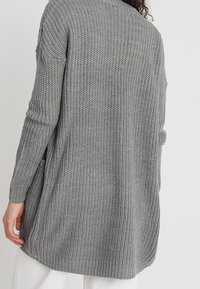 Mujer con un largo y grueso cardigan de punto gris, vista desde atrás, combinado con pantalones blancos sobre un fondo plano.