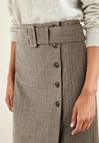 Jupe en tartan marron avec une large ceinture, présentant des fermetures à bouton sur le devant et un tissu texturé. La ceinture a une boucle rectangulaire.