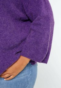 Maglione a maglia viola con finitura testurizzata, caratterizzato da una vestibilità ampia e maniche larghe, abbinato a jeans in denim blu.