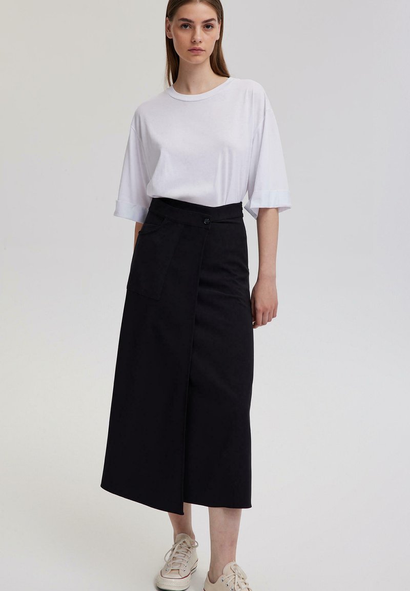 Černá midi sukně s překládaným designem a boční kapsou, spárovaná s volným bílým tričkem. Pohodlné tenisky dokončují outfit.