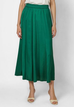 Frau trägt einen hoch taillierten, smaragdgrünen Midi-Rock mit Falten, kombiniert mit beigen offenen Schuhen und einem weißen, in den Rock gesteckten Oberteil.
